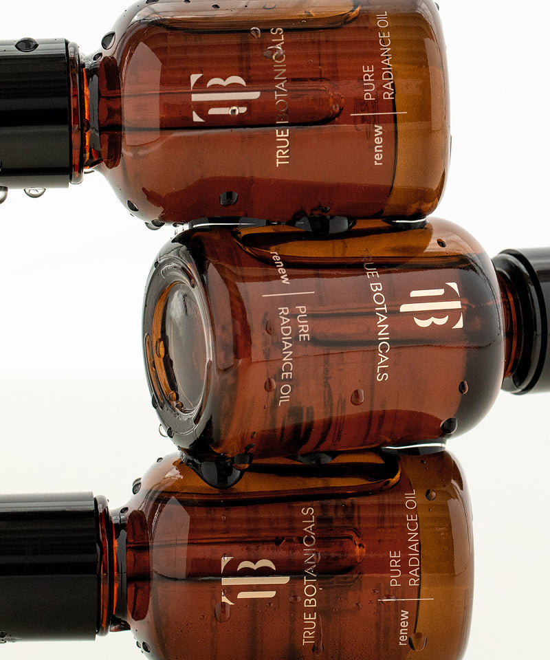 Stacked bottles of True Botanicals Pure Radiance Oil with water droplets, highlighting the product's luxurious and hydrating qualities.