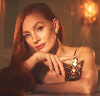 A woman with long, flowing red hair poses with her chin resting on her hand, gazing softly at the camera. She is wearing a black spaghetti-strap top and gold earrings. A small glass skincare bottle reflects light, creating a starburst effect. The background is softly lit with a warm, golden glow and blurred lighting fixtures.