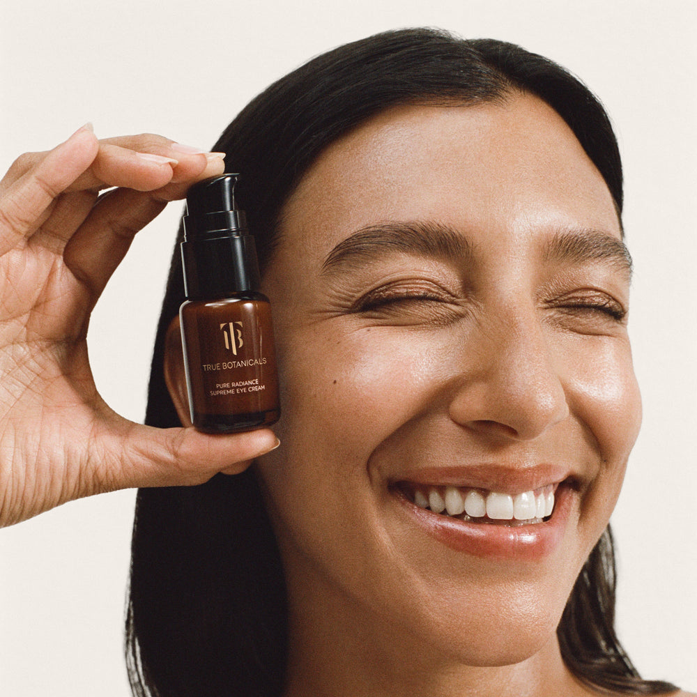 A smiling woman with dark hair holds a small amber pump bottle near her face against a plain light beige background.