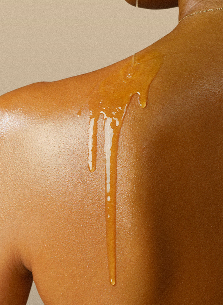 Close-up of a bare shoulder with golden body oil cascading down smooth, glowing skin against a neutral beige background.