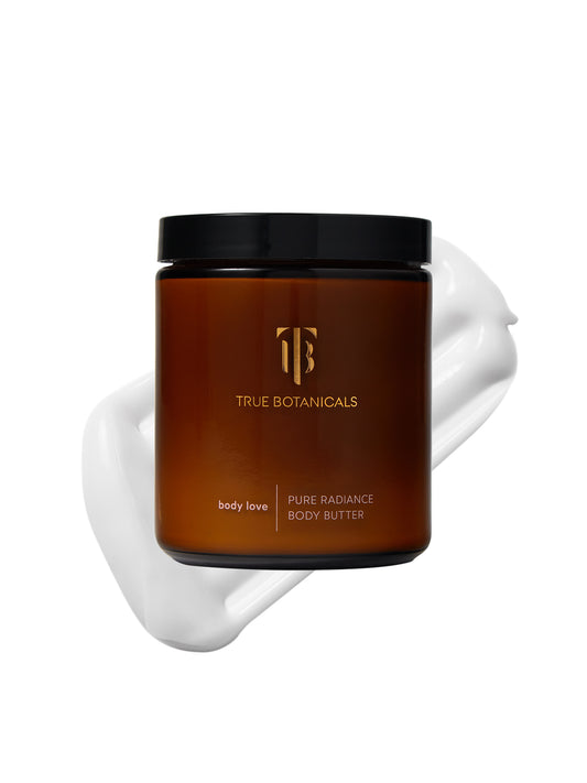 An amber glass jar of True Botanicals Pure Radiance Body Butter with a black lid is displayed against a white background. A smooth, creamy swirl of body butter is positioned behind the jar, highlighting its rich texture. The minimalist label features the True Botanicals logo and product name in gold and white text, conveying a clean and luxurious design.