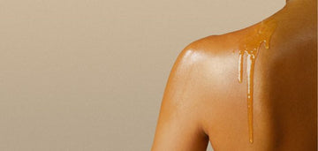 Close-up of a bare shoulder with golden body oil cascading down smooth, glowing skin against a neutral beige background.