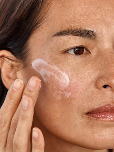 Model with freckles applying True Botanicals Chebula Extreme Cream to their cheek, showing the soft white cream texture being blended into skin against a dark background.