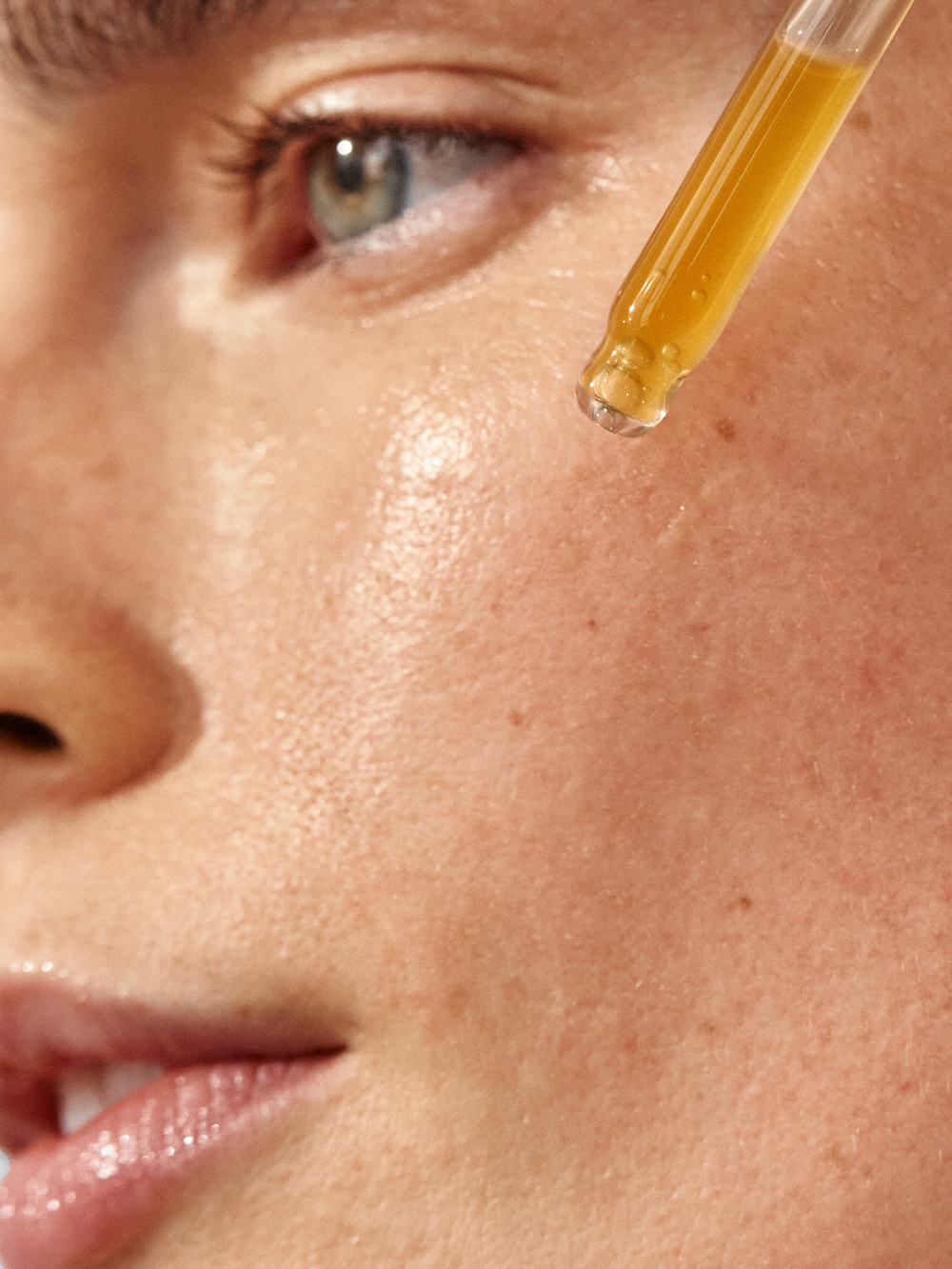 Close-up of a dropper dispensing True Botanicals Chebula Active Serum onto a model's cheek, showing the rich amber-gold serum formula about to be applied to light, freckled skin.