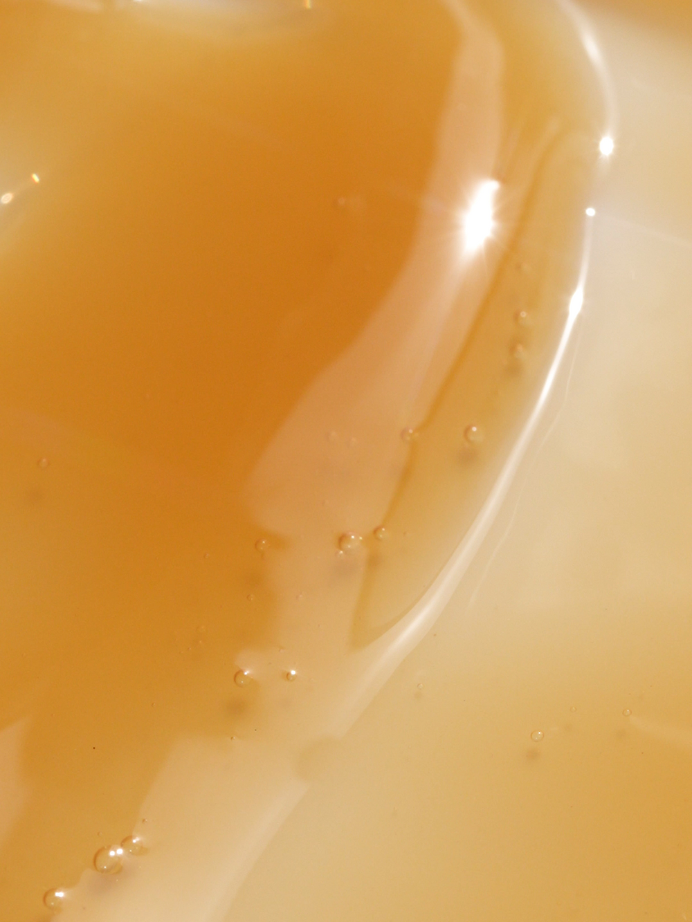 Close-up texture shot of True Botanicals Chebula Active Serum showing a lightweight, fluid amber-gold formula with tiny air bubbles and a glossy, oil-like consistency pooling against a cream-white surface.