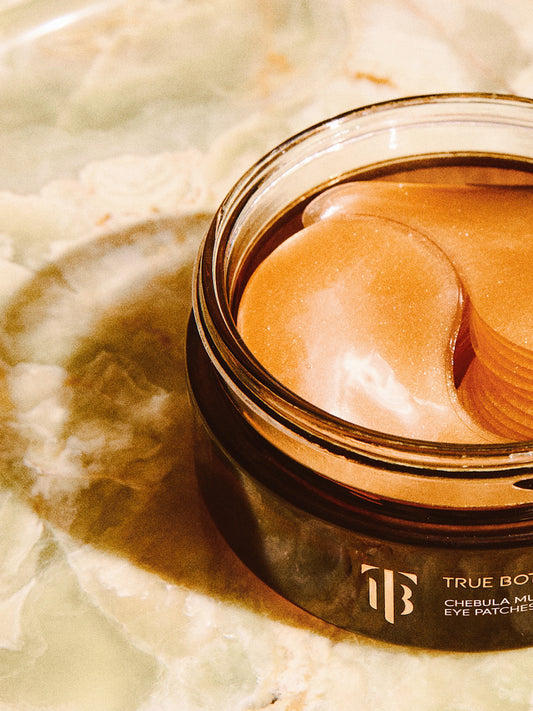 A close-up view of True Botanicals Chebula Multi-Corrective Eye Patches in an elegant amber jar. The shimmering golden patches are arranged inside, displayed on a luxurious marbled surface that enhances the premium aesthetic.