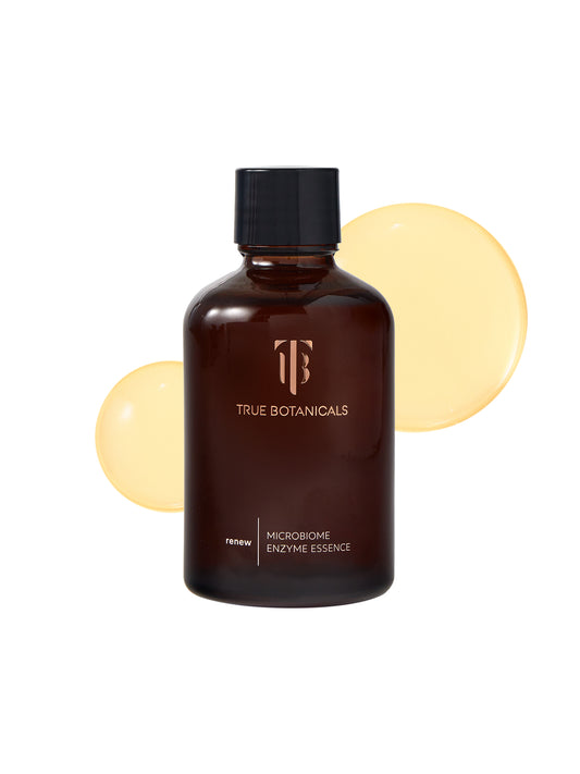 A bottle of True Botanicals Microbiome Enzyme Essence in a dark amber glass container with a black screw cap is displayed against a white background. Behind the bottle, two smooth droplets of golden, translucent liquid are spread, highlighting its lightweight and hydrating formula. The label emphasizes its placement in the "renew" collection.