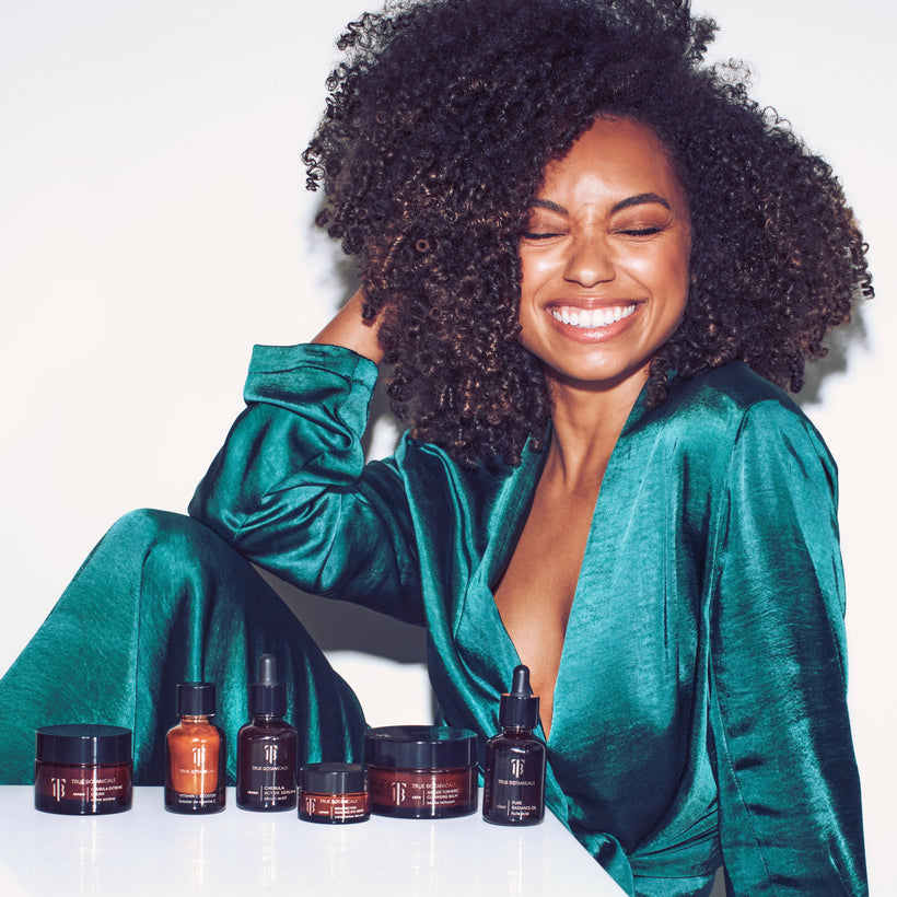 A vibrant image of a smiling woman with glowing skin and natural curls wearing a luxurious emerald green satin outfit. She is sitting next to a collection of True Botanicals skincare products displayed in amber jars and bottles, including creams, serums, and oils. The image exudes confidence, joy, and the essence of radiant self-care. The bright white background enhances the focus on the woman and the products.