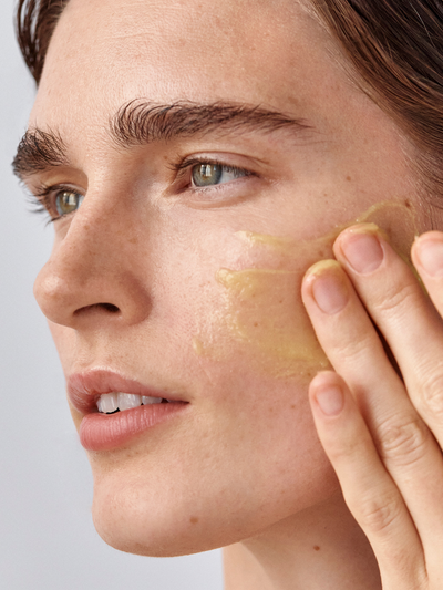 Model with light skin tone applying True Botanicals Calm Ginger Turmeric Cleansing Balm to their cheek, showing the warm golden-yellow balm texture being massaged into skin against a soft gray background.