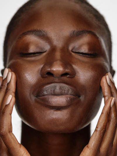 Model with deep skin tone gently pressing both hands to their cheeks with eyes closed, showcasing smooth, glowing skin after using True Botanicals Calm Ginger Turmeric Cleansing Balm against a soft white background.