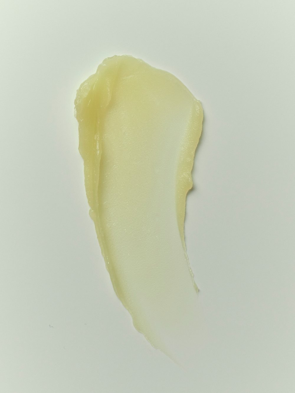 Close-up swatch of True Botanicals Calm Ginger Turmeric Cleansing Balm showing a smooth, glossy pale yellow balm smeared on a light gray surface, with a rich, buttery consistency.