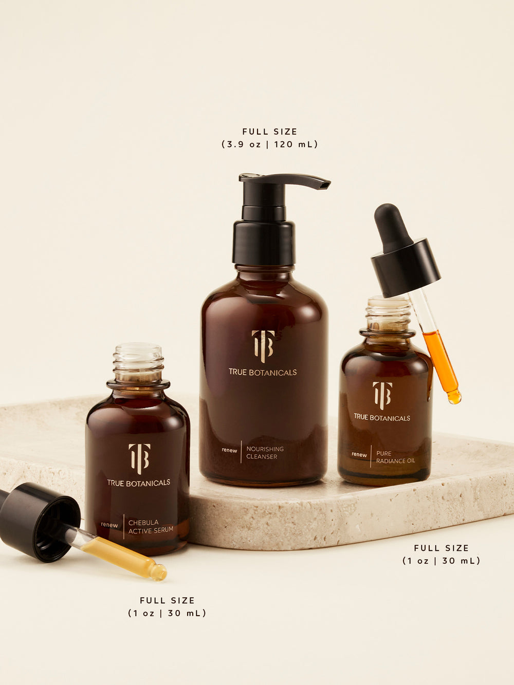 True Botanicals full-size skincare trio — Chebula Active Serum (1 oz | 30 mL), Nourishing Cleanser (3.9 oz | 120 mL), and Pure Radiance Oil (1 oz | 30 mL) — elegantly displayed on a stone platform. Each product is shown open with droppers revealing the golden, nutrient-rich formulas designed to cleanse, hydrate, and visibly renew skin with every step.