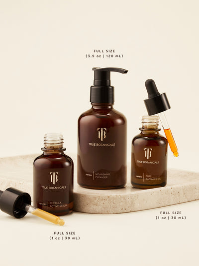 True Botanicals full-size skincare trio — Chebula Active Serum (1 oz | 30 mL), Nourishing Cleanser (3.9 oz | 120 mL), and Pure Radiance Oil (1 oz | 30 mL) — elegantly displayed on a stone platform. Each product is shown open with droppers revealing the golden, nutrient-rich formulas designed to cleanse, hydrate, and visibly renew skin with every step.