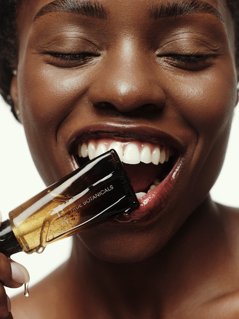 A close-up of a smiling woman holding the True Botanicals Lipid Lip Hydrating Lip Oil tube to her lips, with golden oil dripping from the applicator onto glossy, hydrated lips.