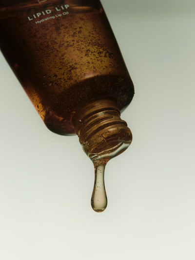 A macro close-up of the True Botanicals Lipid Lip Hydrating Lip Oil tube tilted downward, with a drop of golden oil forming at the applicator tip against a soft grey background.