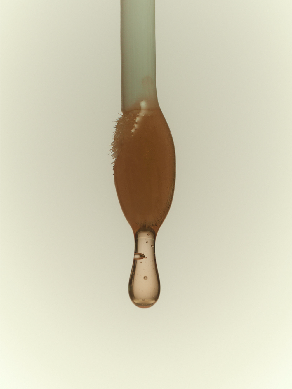 A macro close-up of a dropper coated in warm amber-toned lip oil, with a single translucent drop forming at the tip against a soft cream background.