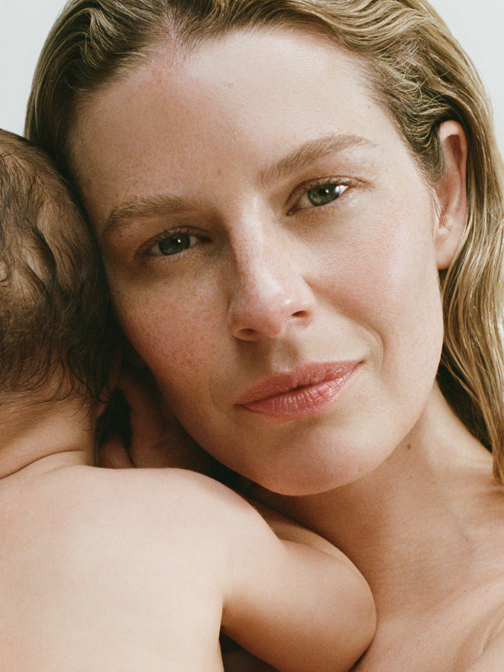 A close-up portrait of a woman holding a baby, with the woman's face and shoulder visible against a soft light background.