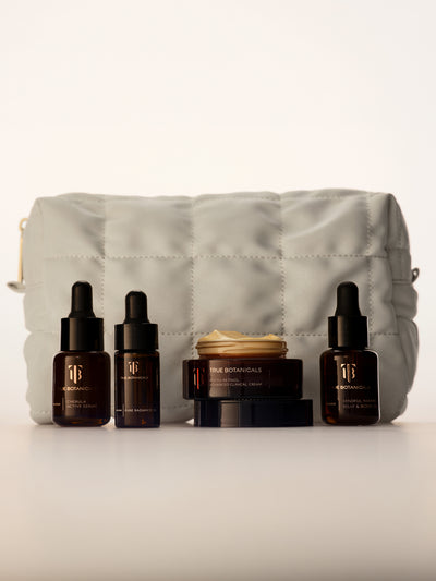A skincare set featuring four amber glass dropper bottles and a jar from True Botanicals, arranged in front of a light gray quilted cosmetic pouch on a clean white background.