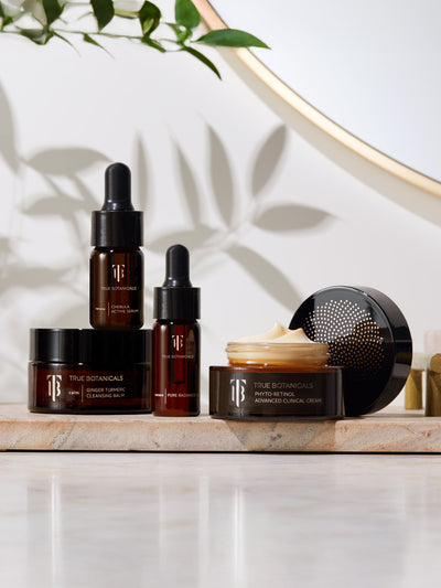 A styled display of True Botanicals skincare products arranged on a natural stone tray. The set includes Chebula Active Serum, Ginger Turmeric Cleansing Balm, Pure Radiance Oil, and Phyto-Retinol Advanced Clinical Cream, all in sleek amber and black packaging. The background features soft plant shadows and a round mirror, creating a luxurious, spa-like atmosphere.