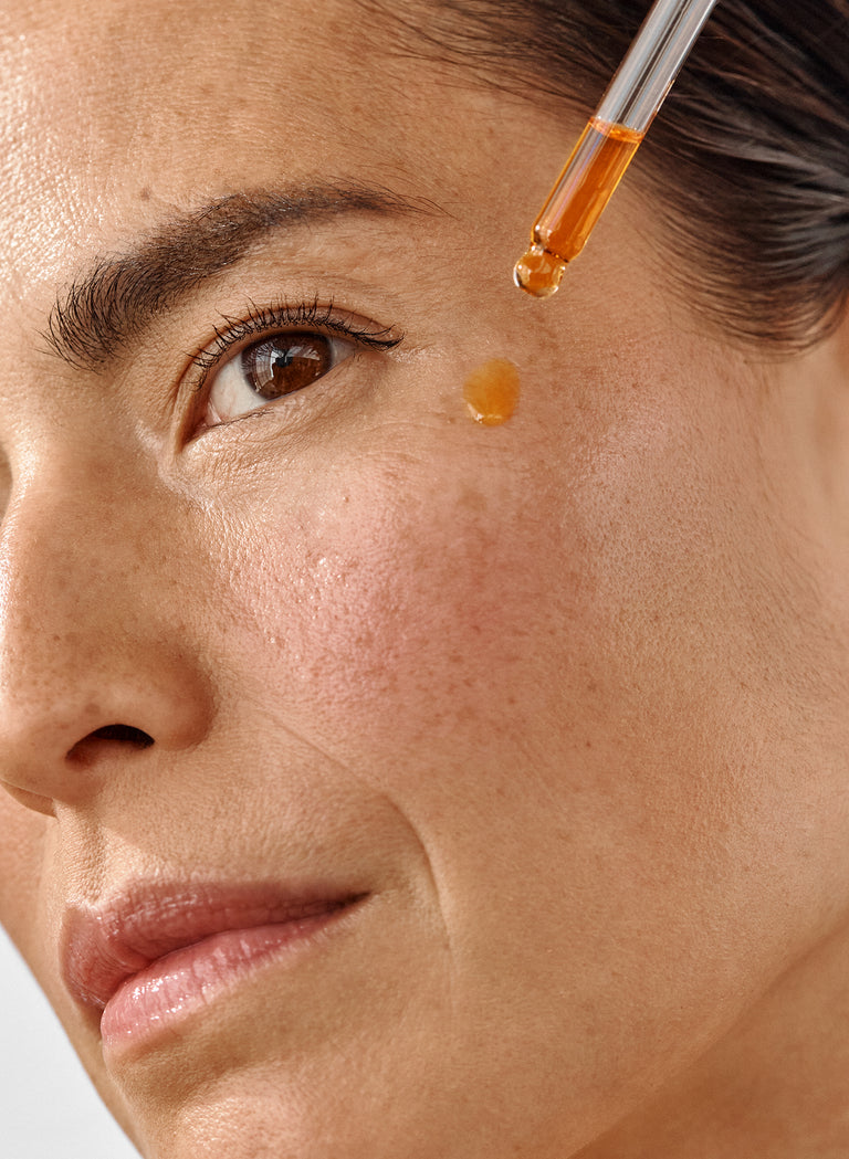 Close-up of a woman's face with a dropper applying golden facial oil onto her cheek, highlighting smooth, radiant skin.