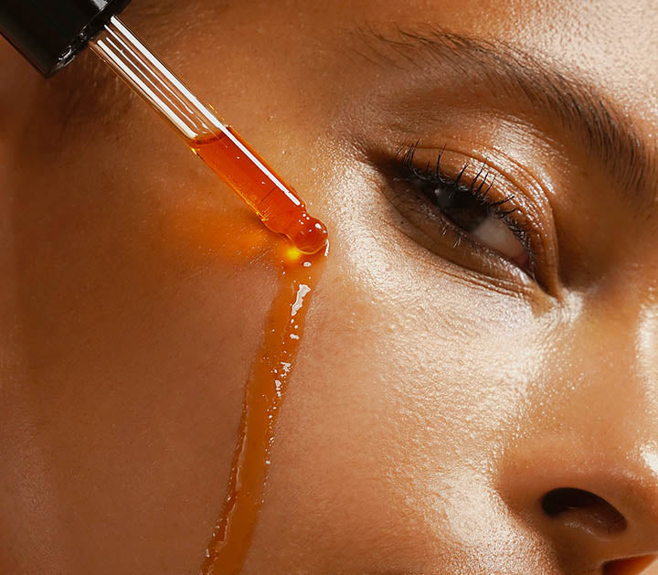 A close-up of a woman’s radiant skin as a dropper applies a golden-hued serum along her cheekbone, highlighting hydration and glow. The image emphasizes a luxurious skincare experience.