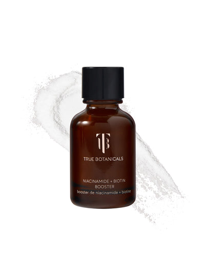 A bottle of True Botanicals Niacinamide + Biotin Booster in a dark amber glass container with a black screw cap is displayed against a white background. Behind the bottle, a swirl of fine, white powder is spread, emphasizing its concentrated, powdered formula. The label highlights its key ingredients, niacinamide and biotin.