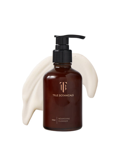 A bottle of True Botanicals Clear Nourishing Cleanser in a dark amber glass container with a black pump dispenser is displayed against a white background. Behind the bottle, a smooth swirl of creamy, white cleanser is spread, highlighting its rich and gentle texture. The label emphasizes its placement in the "clear" collection.