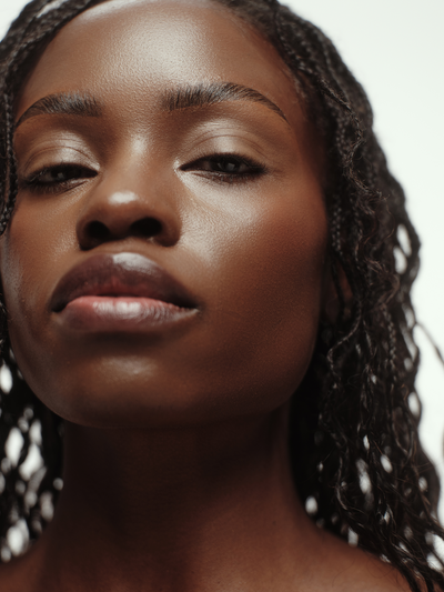 Model with deep skin tone and braided hair gazing upward, showcasing an exceptionally smooth, luminous complexion after using True Botanicals Renew Pure Radiance Oil against a soft white background.
