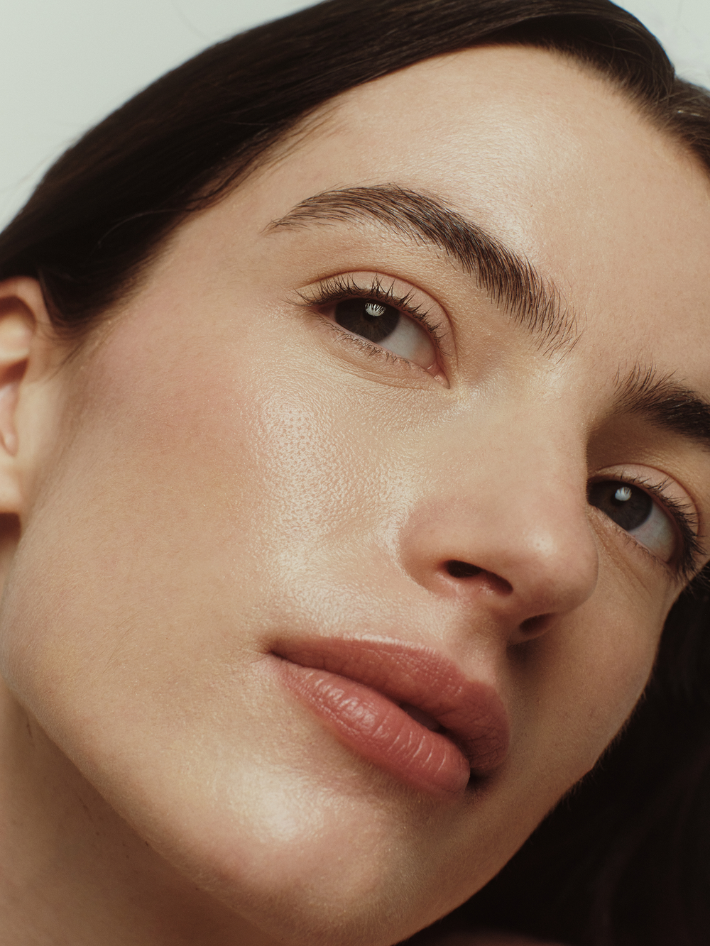 Model with light skin tone and dark hair looking upward, showcasing a dewy, glass-skin complexion with a visible lit-from-within glow after using True Botanicals Renew Pure Radiance Oil against a soft neutral background.