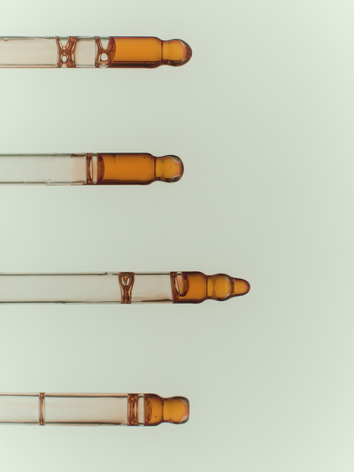 Four glass dropper pipettes filled with True Botanicals Renew Pure Radiance Oil arranged diagonally, showing the rich amber-gold oil formula inside each glass tube against a soft sage-green background.