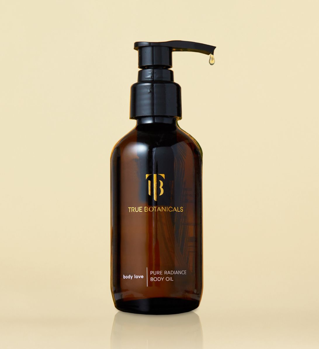 Amber glass bottle of True Botanicals Pure Radiance Body Oil with a black pump dispenser and a droplet of oil at the tip, set against a soft beige background.