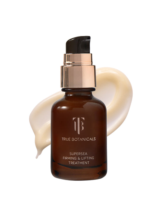 A bottle of True Botanicals Supersea Firming & Lifting Treatment in a dark amber glass container with a pump dispenser is displayed against a white background. Behind the bottle, a smooth swirl of creamy, off-white serum is visible.