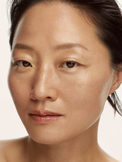 A close-up portrait of a woman with natural skin texture and makeup, showing her face from the shoulders up against a plain light background.