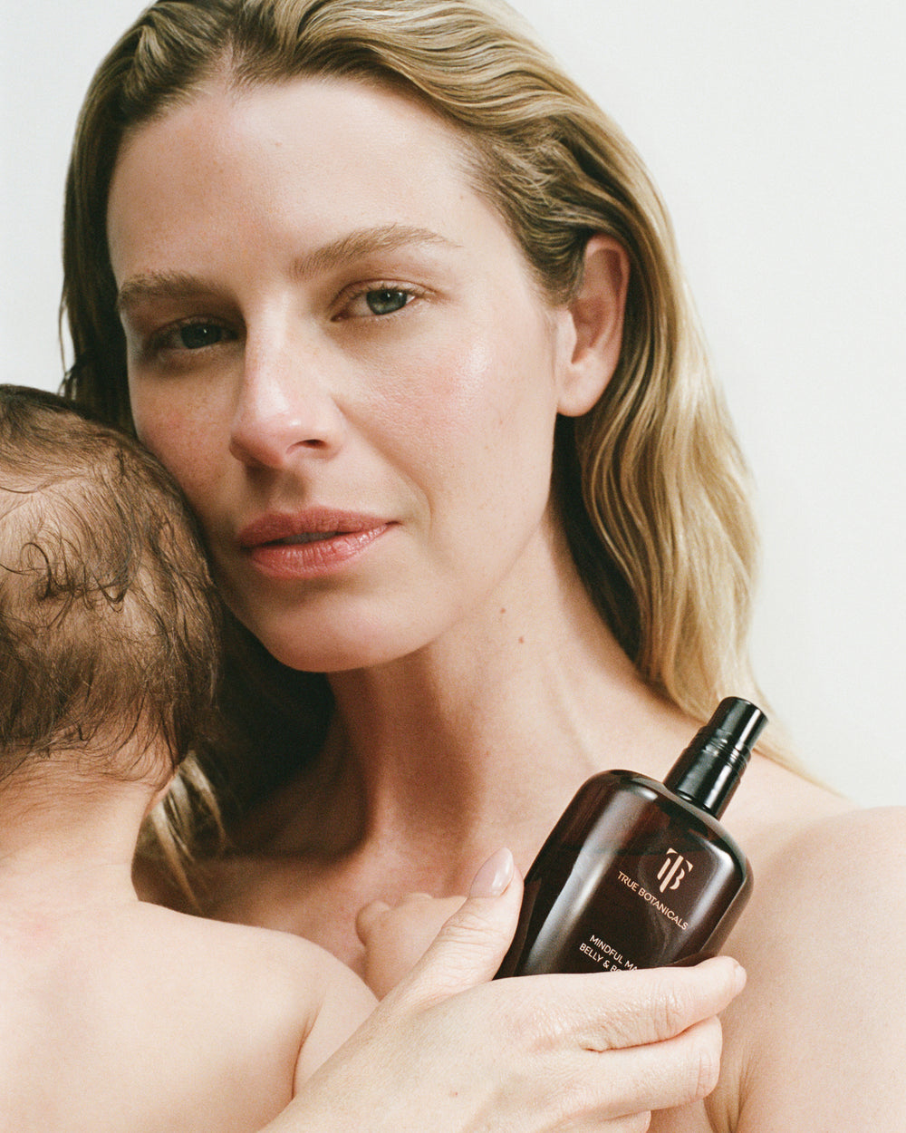 A woman holding a dark brown bottle of Mindful Mama Belly and body oil while embracing a baby against a clean white background.