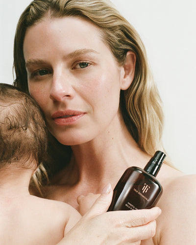 A woman holding a dark brown bottle of Mindful Mama Belly and body oil while embracing a baby against a clean white background.
