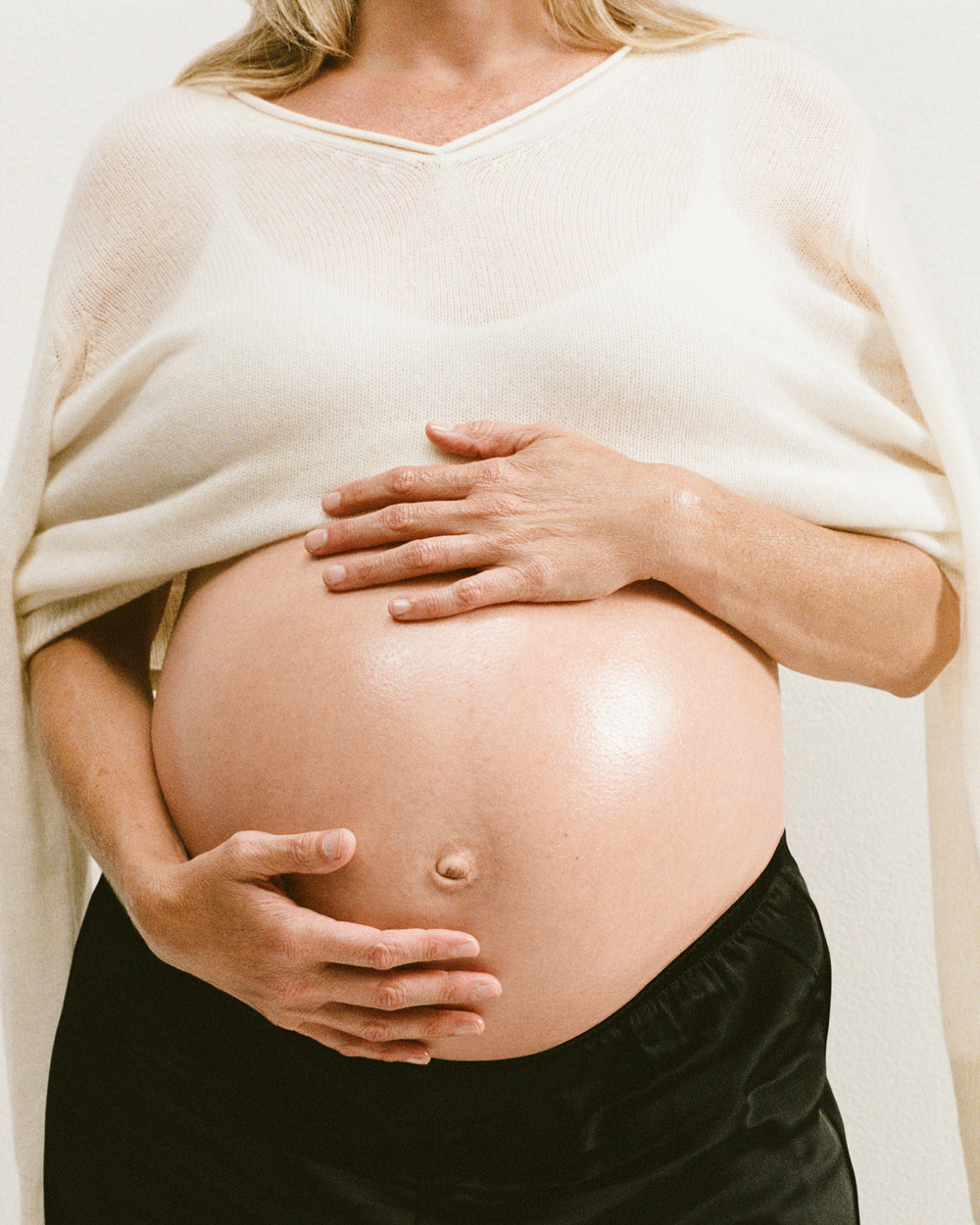 Pregnant woman gently cradling her belly with both hands, wearing a soft cream top and black pants.