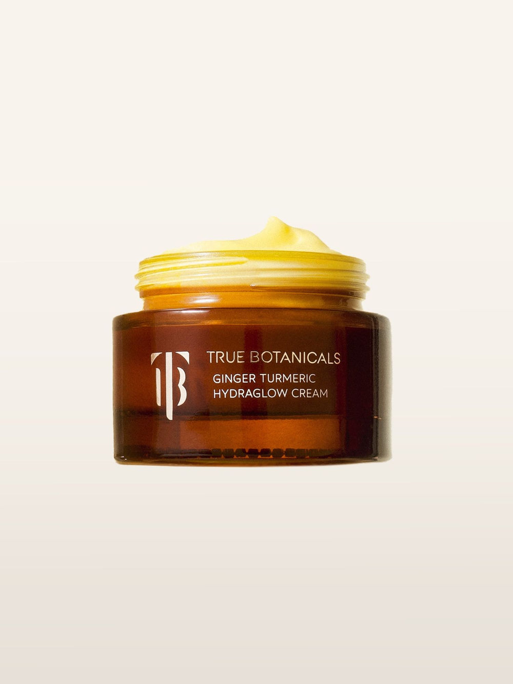 The image highlights True Botanicals' Ginger Turmeric Hydraglow Cream, showcasing its luxurious and creamy texture in an amber glass jar. The minimalist background emphasizes the product’s golden hue and rich formulation, perfect for conveying the essence of natural glow and hydration.