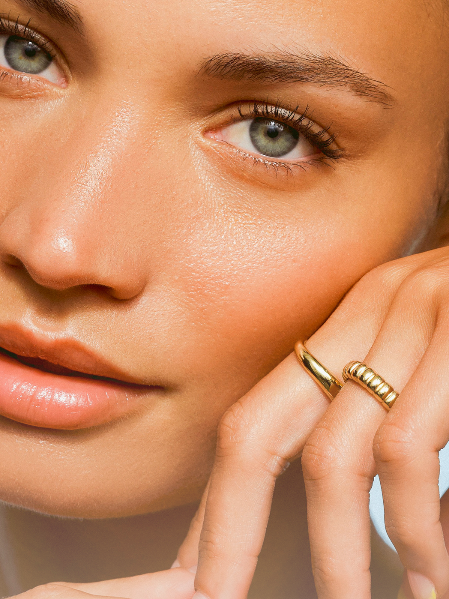 Close-up of a woman with luminous, hydrated skin and green eyes, gently touching her face, wearing gold rings on her fingers.